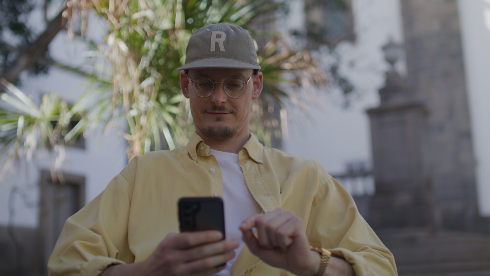 Mężczyzna w żółtej koszuli i okularach przegląda telefon na zewnątrz, nosząc czapkę z literą „R” i stojąc w otoczeniu palm.