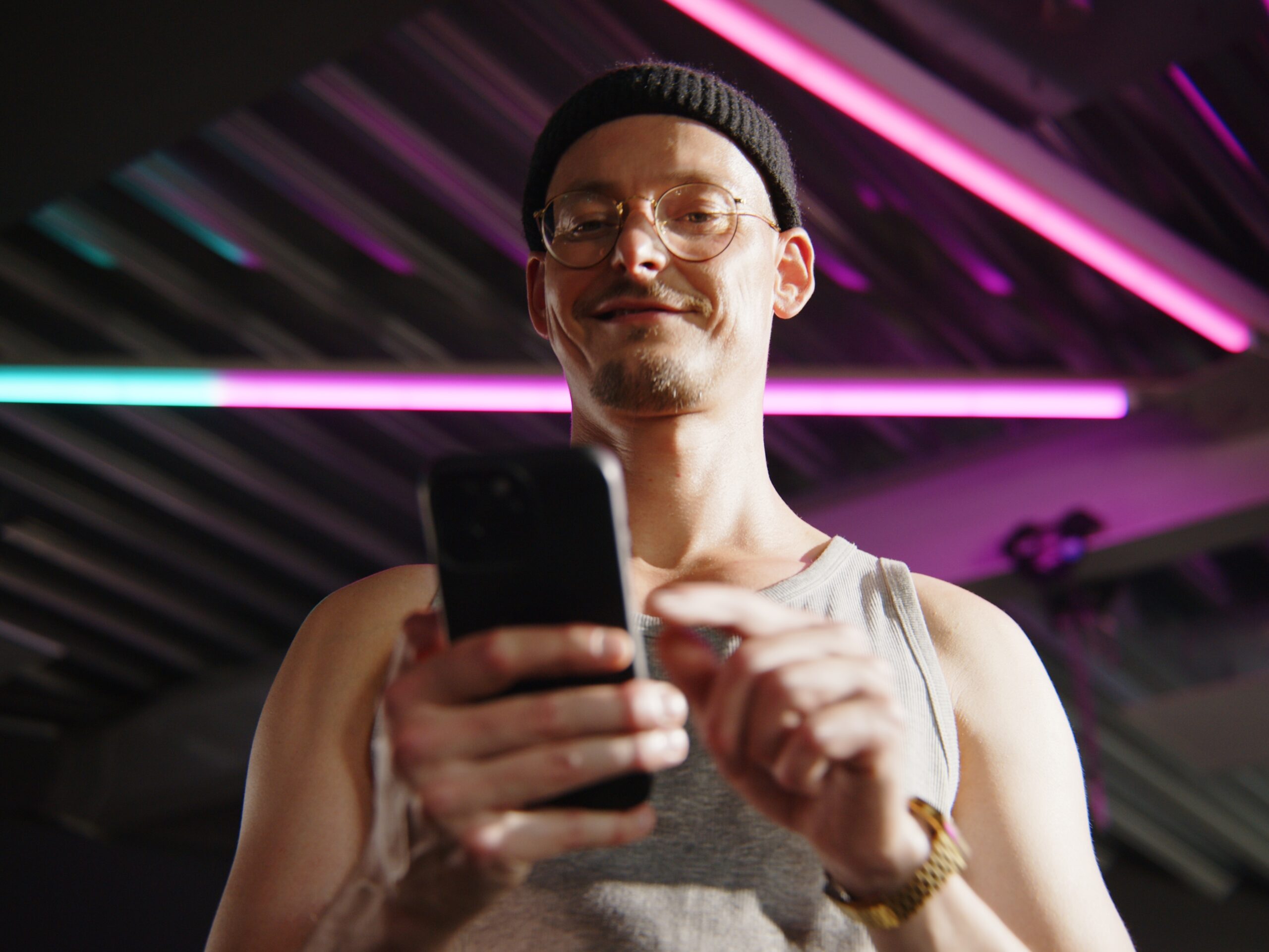 Mężczyzna w czapce i okularach trzyma smartfona i uśmiecha się, stojąc pod neonowymi kolorowymi światłami.